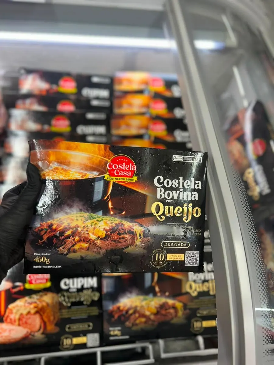 Mão segurando uma caixa de Costela Bovina com Queijo na frente do freezer de um supermercado